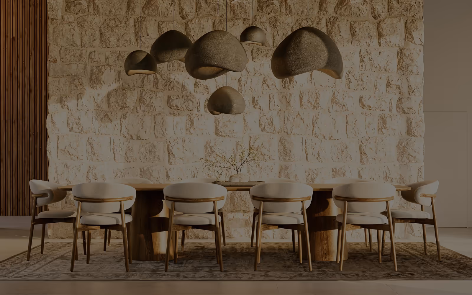 Mesa de jantar comprida em madeira com cadeiras estofadas claras, sob sete candeeiros suspensos de formas orgânicas, diante de uma parede de pedra aparelhada.