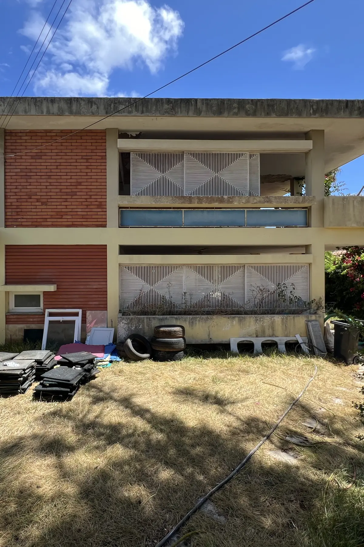 O alçado do jardim de frente: pano de tijolo à esquerda, dois pisos recuados com painéis vazados brancos e uma faixa azul, a laje de cobertura a avançar por cima, e materiais encostados ao muro da base.