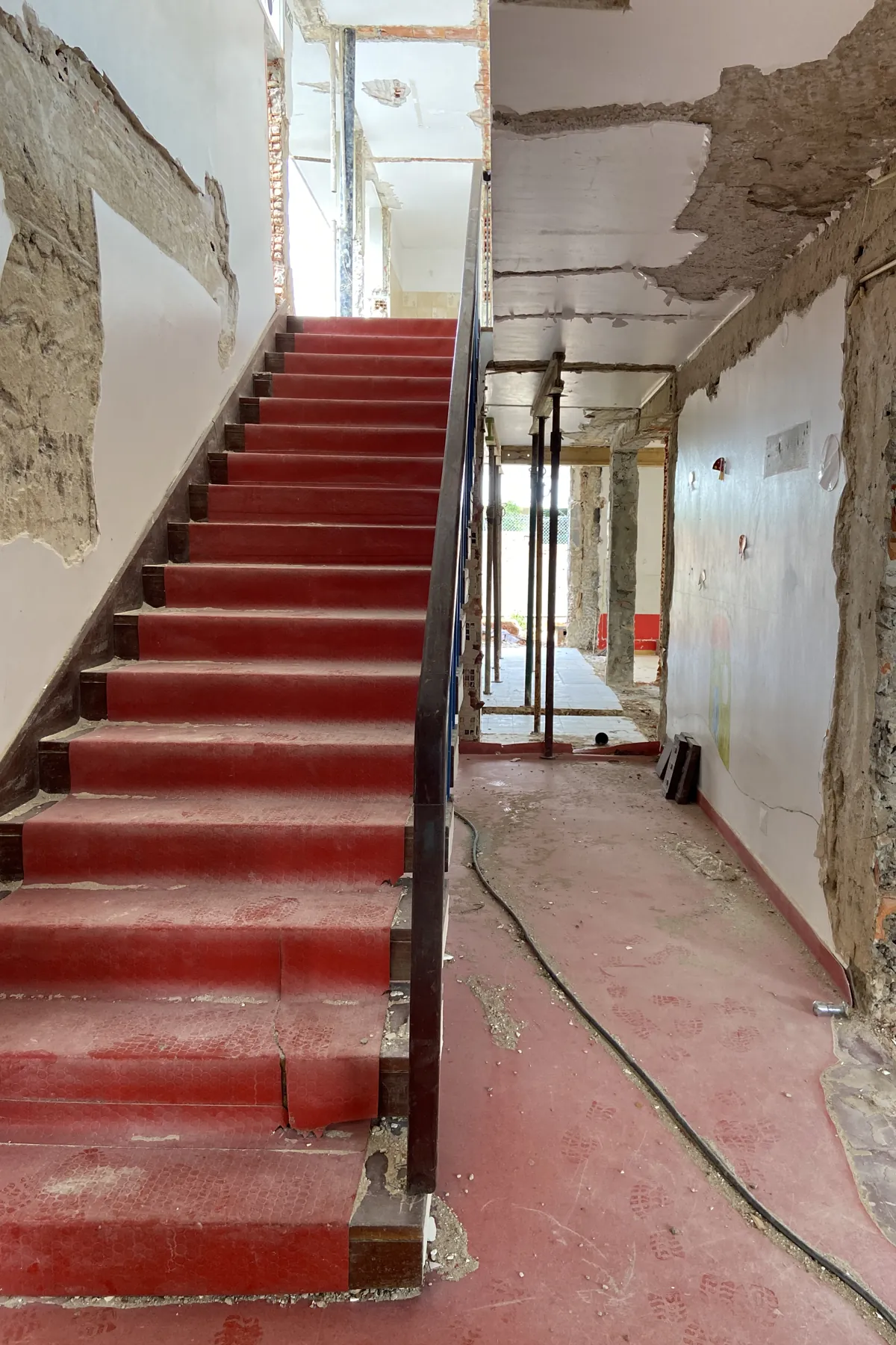 Um lanço de escada direito acabado a vermelho, a subir contra uma parede com o revestimento retirado, com corrimão metálico escuro e, ao fundo, um corredor com prumos.