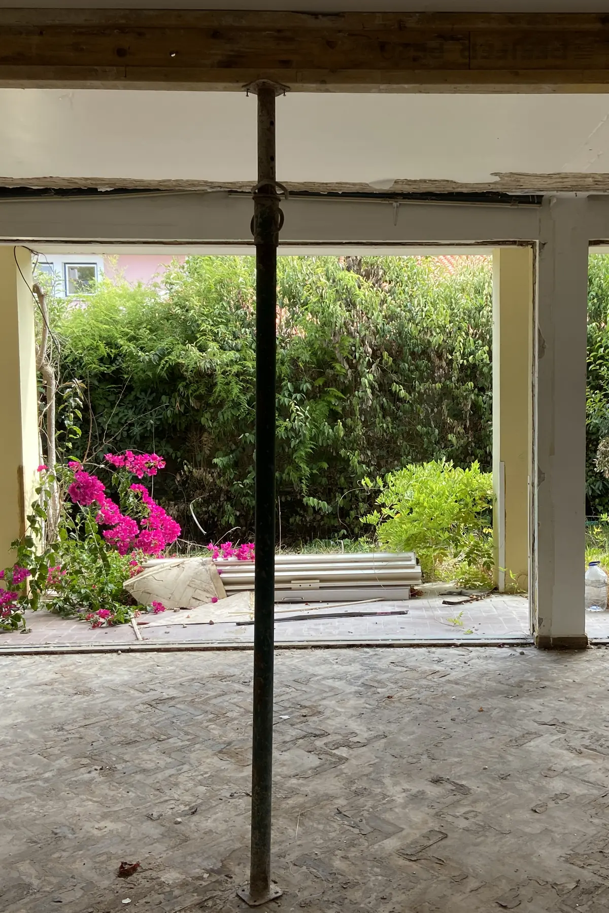 Uma sala do piso térreo com as divisórias retiradas, um prumo metálico sob a viga, e a parede aberta para o jardim, com buganvília em flor contra a sebe.