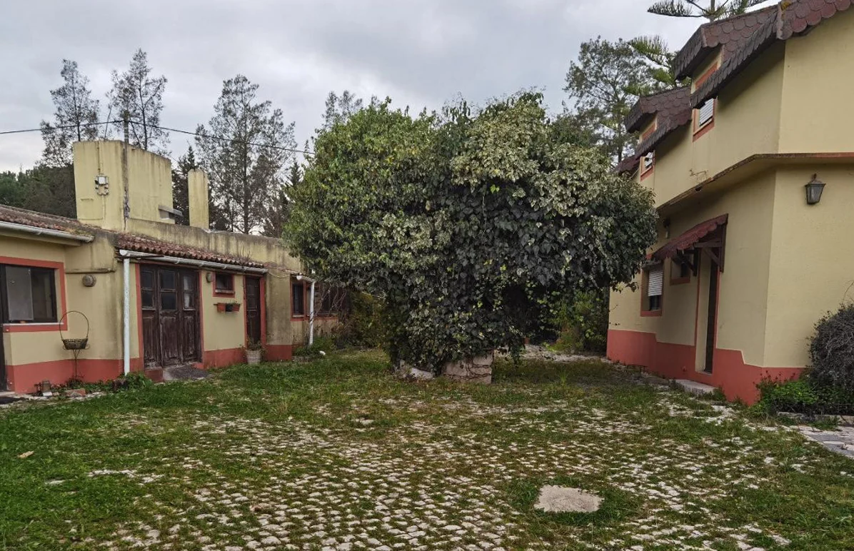Um pátio entre edifícios térreos rebocados de amarelo claro com embasamentos pintados de encarnado, um relvado por tratar e um caminho de calçada, e ao fundo uma casa mais alta com telhado de telha.