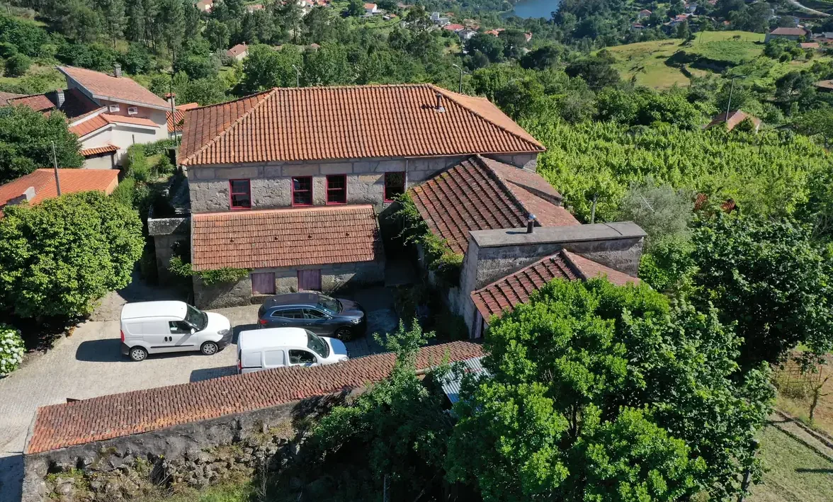 Uma vista aérea de um solar de granito com telhados de telha entre vinha e mato, com um vale de rio atrás.