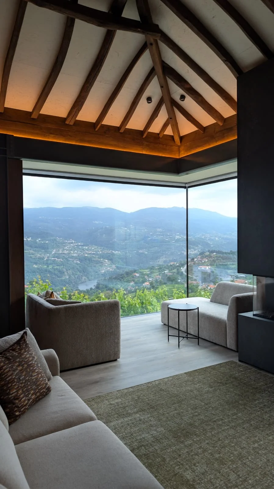 Uma sala de estar acabada sob a cobertura de madeira, com um canto de vidro de pé-direito completo sobre o vale, sofás baixos, uma mesa de apoio redonda e um tapete verde.