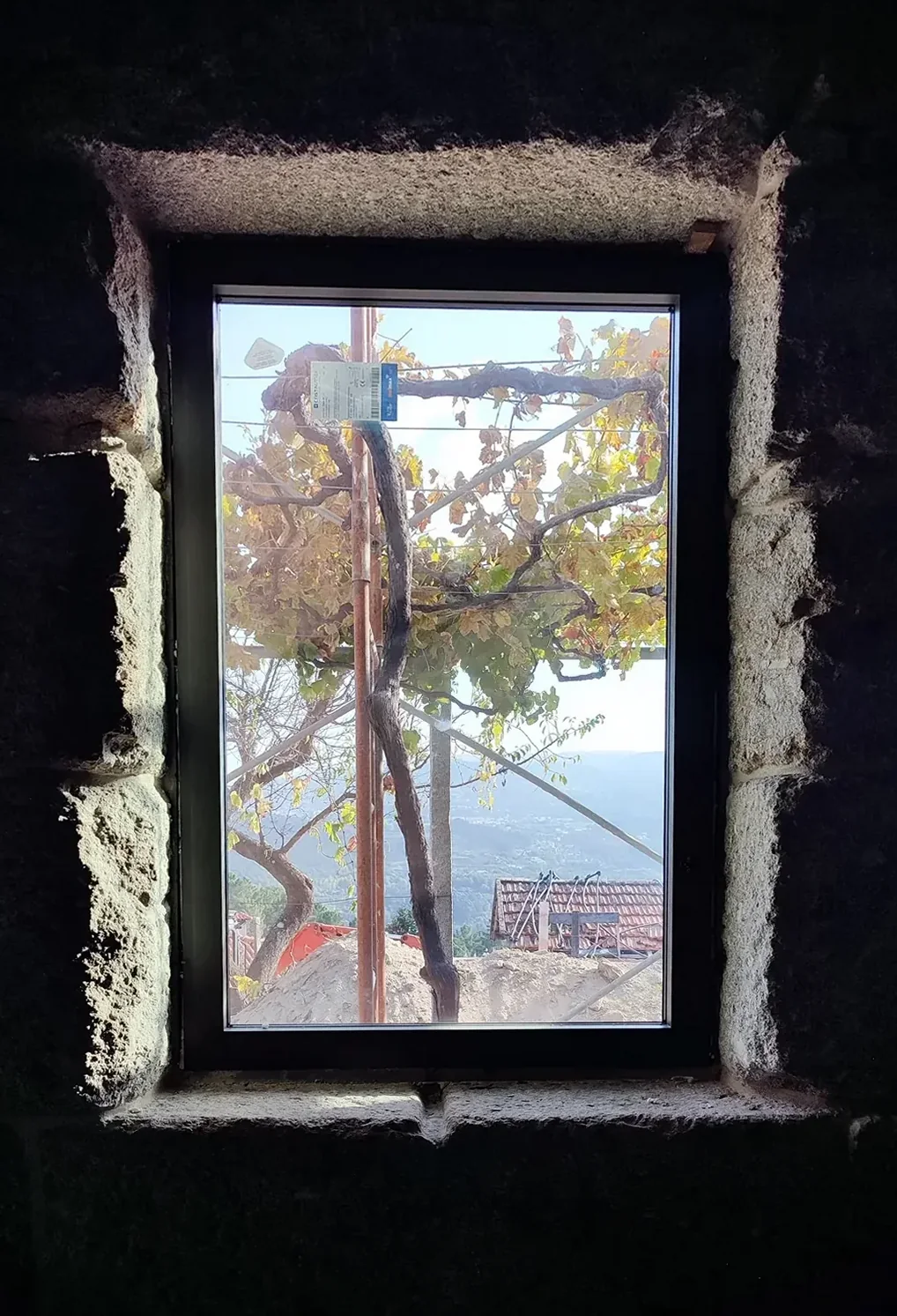 Uma janela encaixada fundo numa parede de granito escura, a enquadrar o tronco de uma videira e o vale ao fundo.