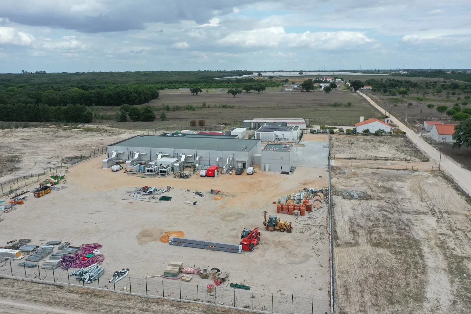 Uma vista aérea do conjunto acabado: edifícios brancos, um estaleiro vedado e pavimentado com máquinas e equipamento, e campo aberto em volta.