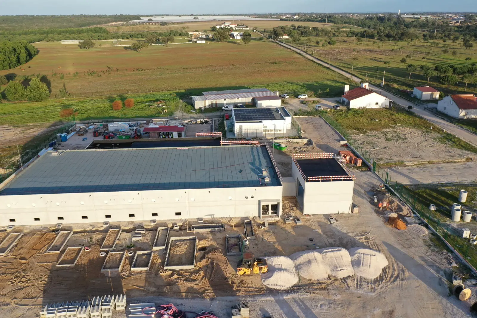 Uma vista aérea de um edifício de produção comprido e branco com cobertura metálica, concluído, com um segundo edifício menor ao lado e sapatas ainda em trabalho em primeiro plano.