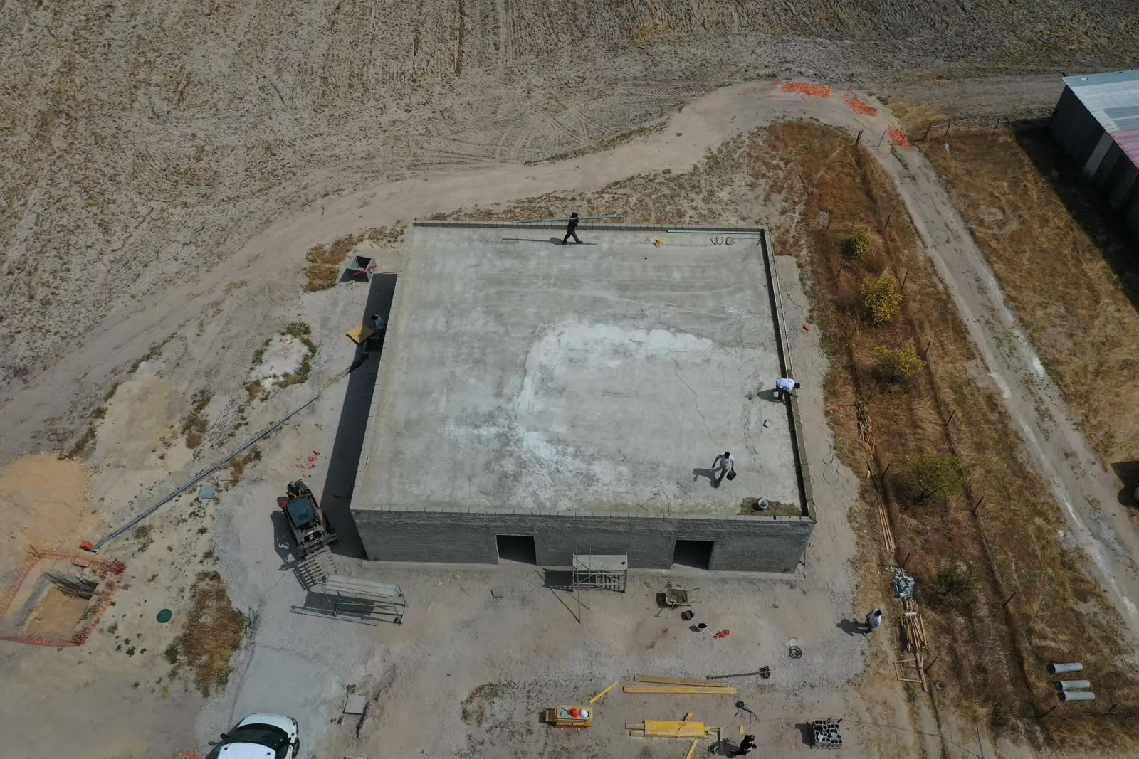 Uma vista aérea de um único invólucro de betão com a laje de cobertura betonada, sozinho numa plataforma limpa com máquinas e materiais em volta.