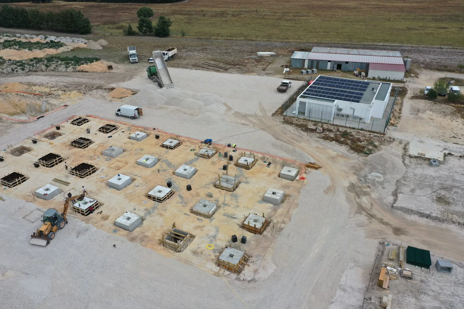 Uma vista aérea a mostrar um edifício branco concluído com painéis solares na cobertura ao lado de uma malha de sapatas recém-betonadas dispostas numa plataforma limpa.