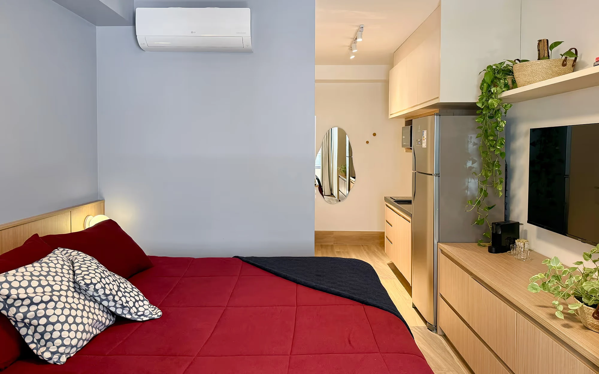 O studio visto da cama: colcha vermelha, espelho oval no corredor, cozinha pequena com geladeira, televisão e móvel de madeira com plantas., Apartamento Cayowaá, Vila Madalena, São Paulo