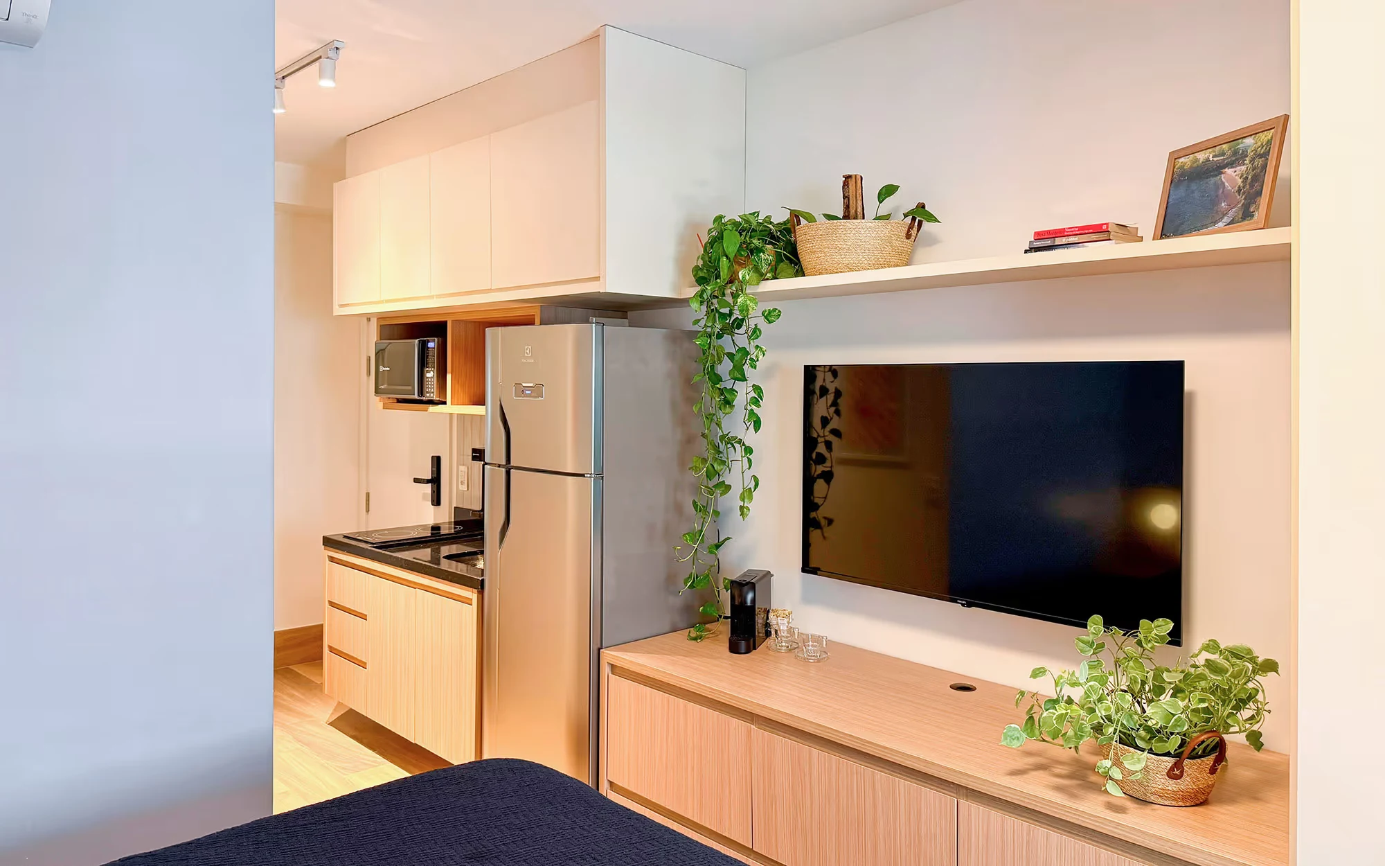 Studio com cozinha pequena, geladeira prateada e armários claros, televisão sobre um móvel de madeira clara e prateleira com plantas, com a cama em primeiro plano., Apartamento Cayowaá, Vila Madalena, São Paulo