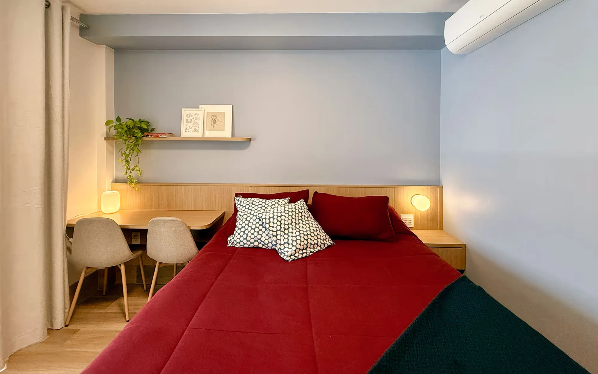 Quarto de paredes azul-claras com cama de colcha vermelha, cabeceira de madeira com arandelas, escrivaninha com duas cadeiras e prateleira com quadros e uma planta., Apartamento Cayowaá, Vila Madalena, São Paulo