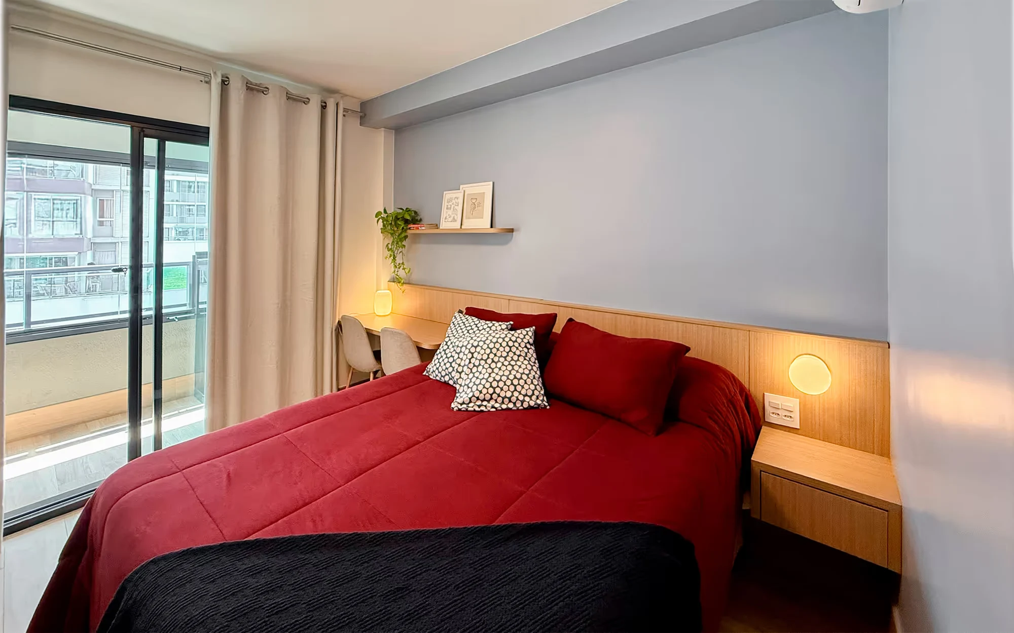 Quarto com colcha vermelha e manta preta, cabeceira de madeira com luminárias redondas, escrivaninha junto à cortina e porta de vidro para a varanda, com prédios lá fora., Apartamento Cayowaá, Vila Madalena, São Paulo