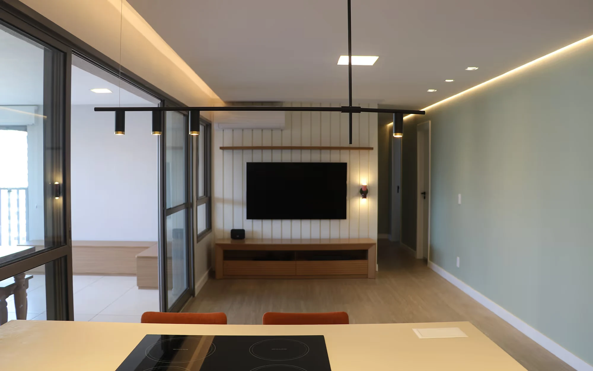 Da cozinha para a sala: televisão em um painel de ripas verticais, rack baixo de madeira, parede verde-clara com luz indireta e portas de correr para a varanda., Apartamento Tucuna, Perdizes, São Paulo