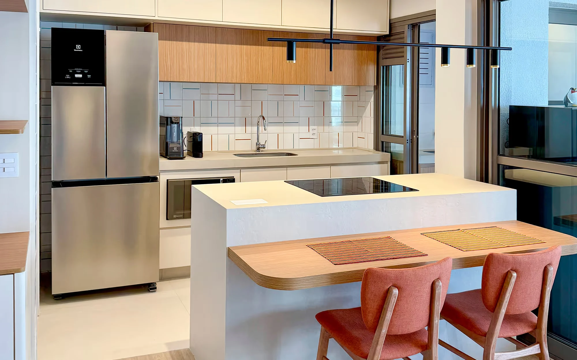 Cozinha compacta com geladeira prateada, armários claros e de madeira, ilha com cooktop preto e balcão curvo de madeira com duas cadeiras cor de terracota., Apartamento Tucuna, Perdizes, São Paulo