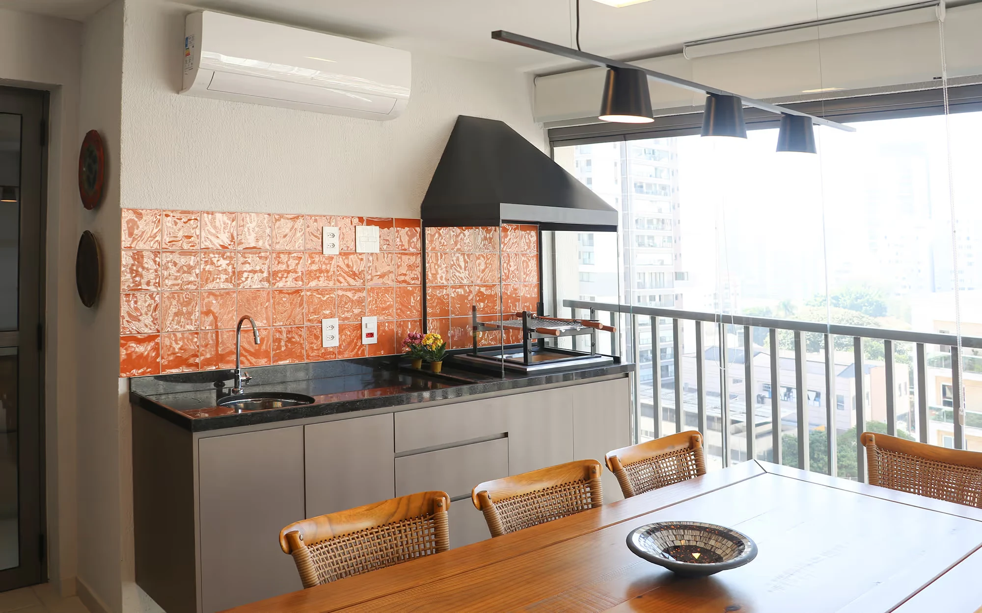 Varanda gourmet com azulejo laranja metalizado, pia e churrasqueira, e uma mesa com cadeiras de palhinha sob três pendentes pretos, com prédios lá fora., Apartamento Tucuna, Perdizes, São Paulo