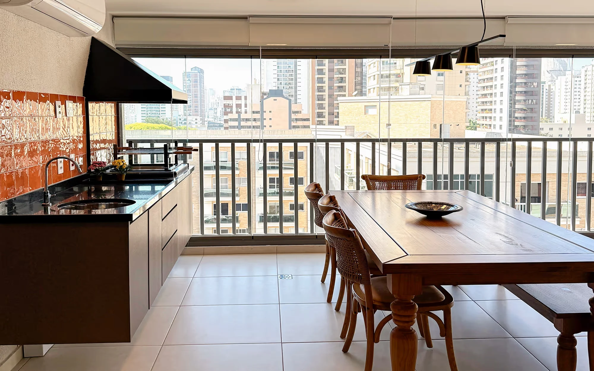 Varanda envidraçada com bancada comprida, pia e churrasqueira, mesa de madeira com cadeiras de palhinha e um banco, e prédios altos atrás do vidro., Apartamento Tucuna, Perdizes, São Paulo