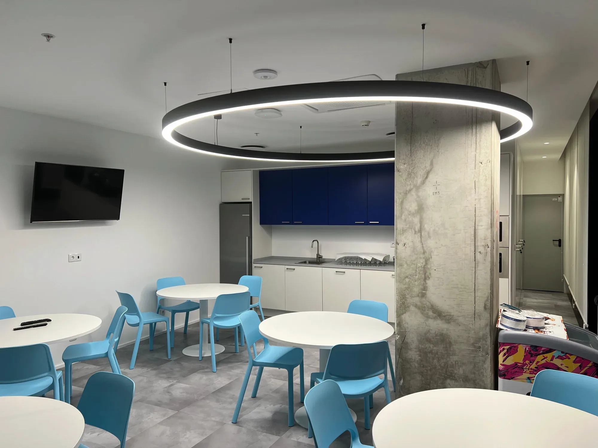 Sala de refeições com mesas redondas brancas e cadeiras moldadas azul-claras sobre pavimento cerâmico cinzento, uma copa branca e azul-escura ao fundo, e uma grande luminária em anel preta ao lado de um pilar de betão à vista.
