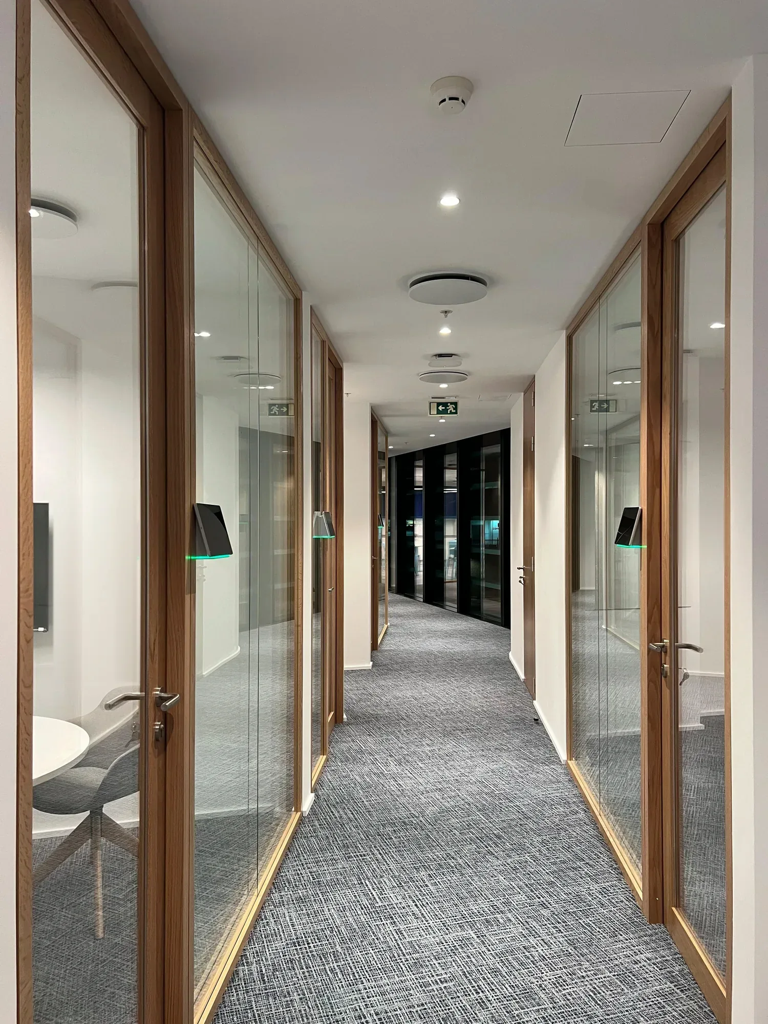 Corredor entre duas filas de salas de reunião em vidro transparente com caixilharia de carvalho, com um painel de reserva iluminado ao lado de cada porta, alcatifa cinzenta texturada, e uma parede envidraçada escura a fechar a vista ao fundo.