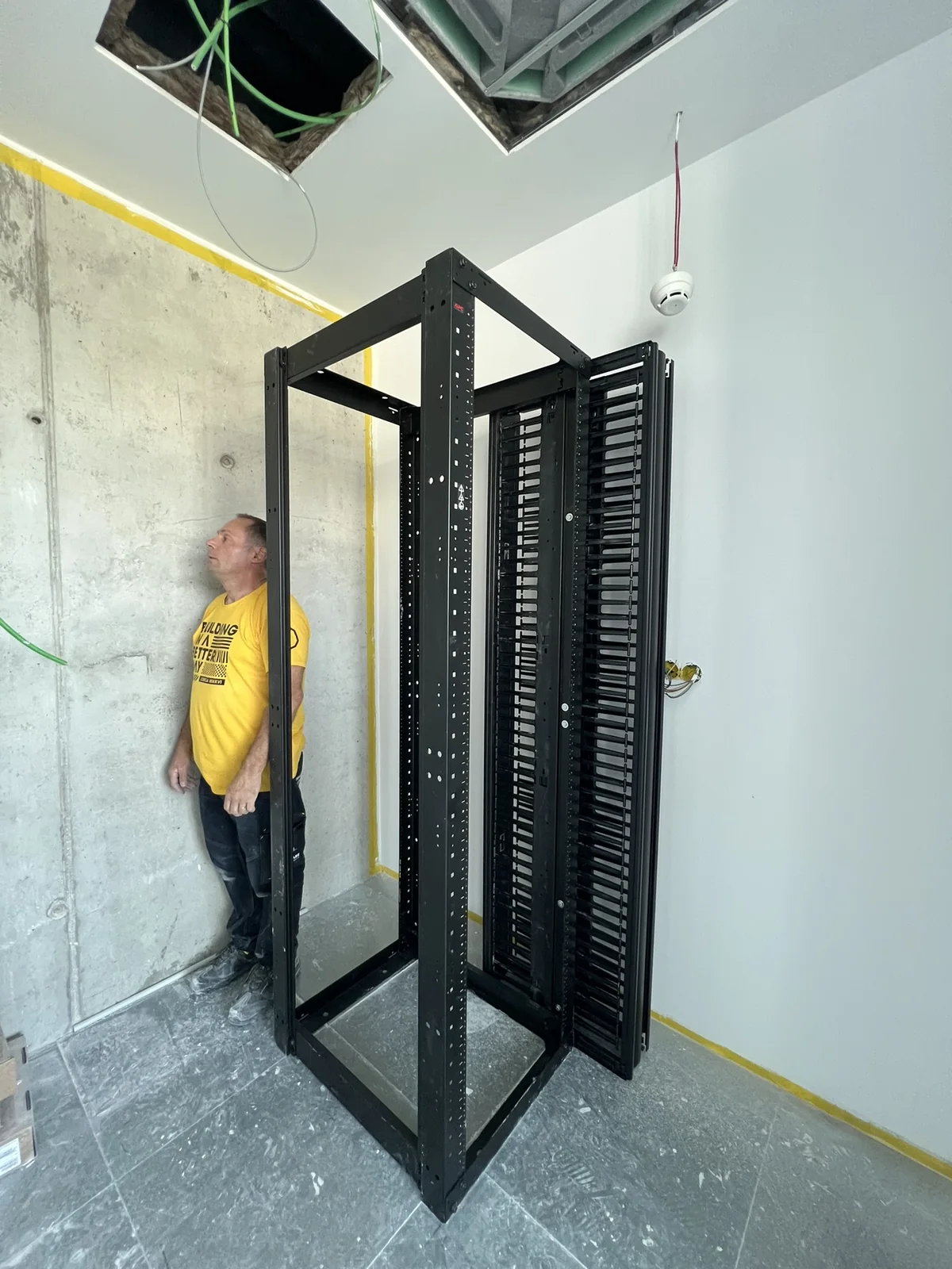 A estrutura do bastidor por montar, no meio da sala que o vai receber, com as portas de lamelas encostadas ao lado; o electricista está junto à parede a ver o espaço que sobra à volta.