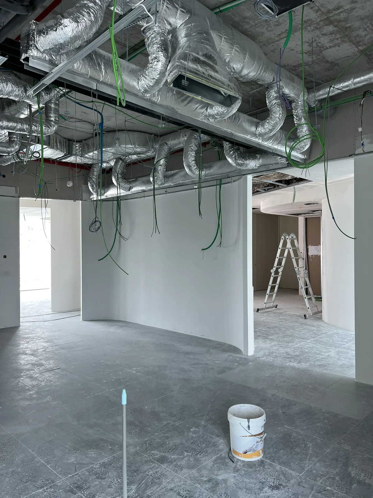 Condutas de AVAC isoladas a correr num tecto aberto, com cablagem descida e um escadote encostado a uma divisória.