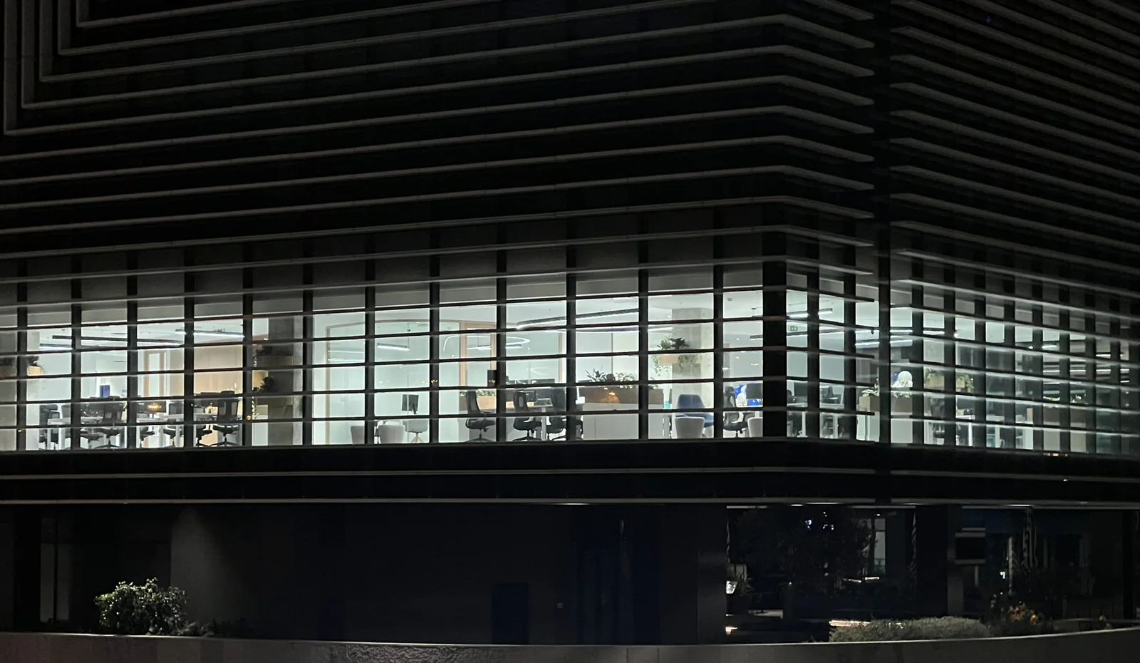 O edifício de escritórios visto da rua, de noite, com o brise-soleil horizontal escuro contra o céu e um piso aceso por dentro: filas de cadeiras vazias, secretárias e salas envidraçadas visíveis através do envidraçado.