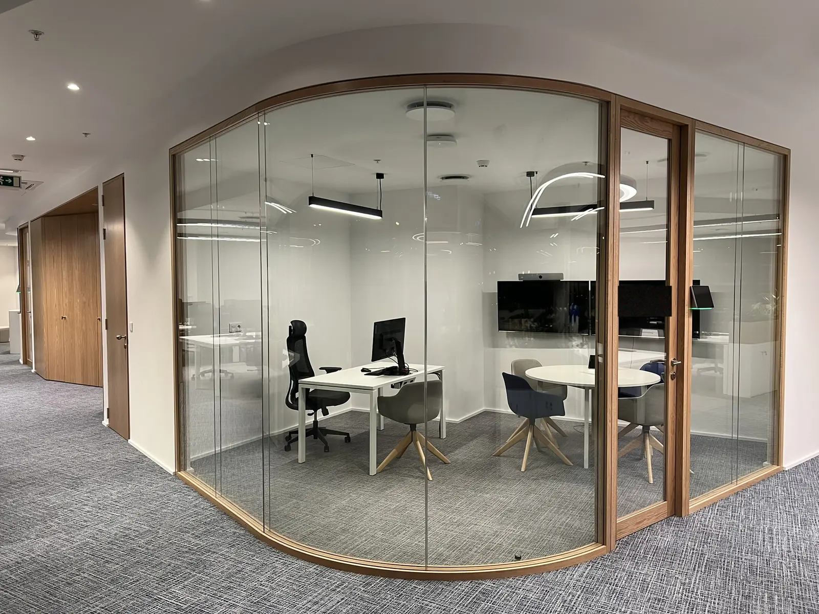 Sala de reunião fechada por uma única parede de vidro curvo em caixilho de carvalho claro, com a planta inteira visível do exterior: secretária, ecrã, mesa redonda e quatro cadeiras, com um candeeiro linear dentro e um anel ao fundo.