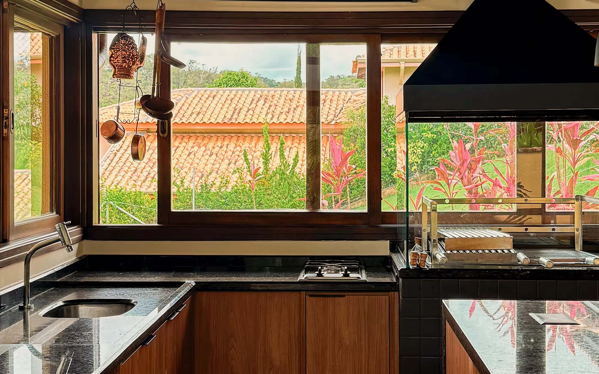 Bancada de tampo escuro sob uma janela larga, com pia, cooktop e churrasqueira sob coifa preta, panelas penduradas e telhados de telha lá fora., Casa Flamboyant Gourmet, Valinhos, São Paulo