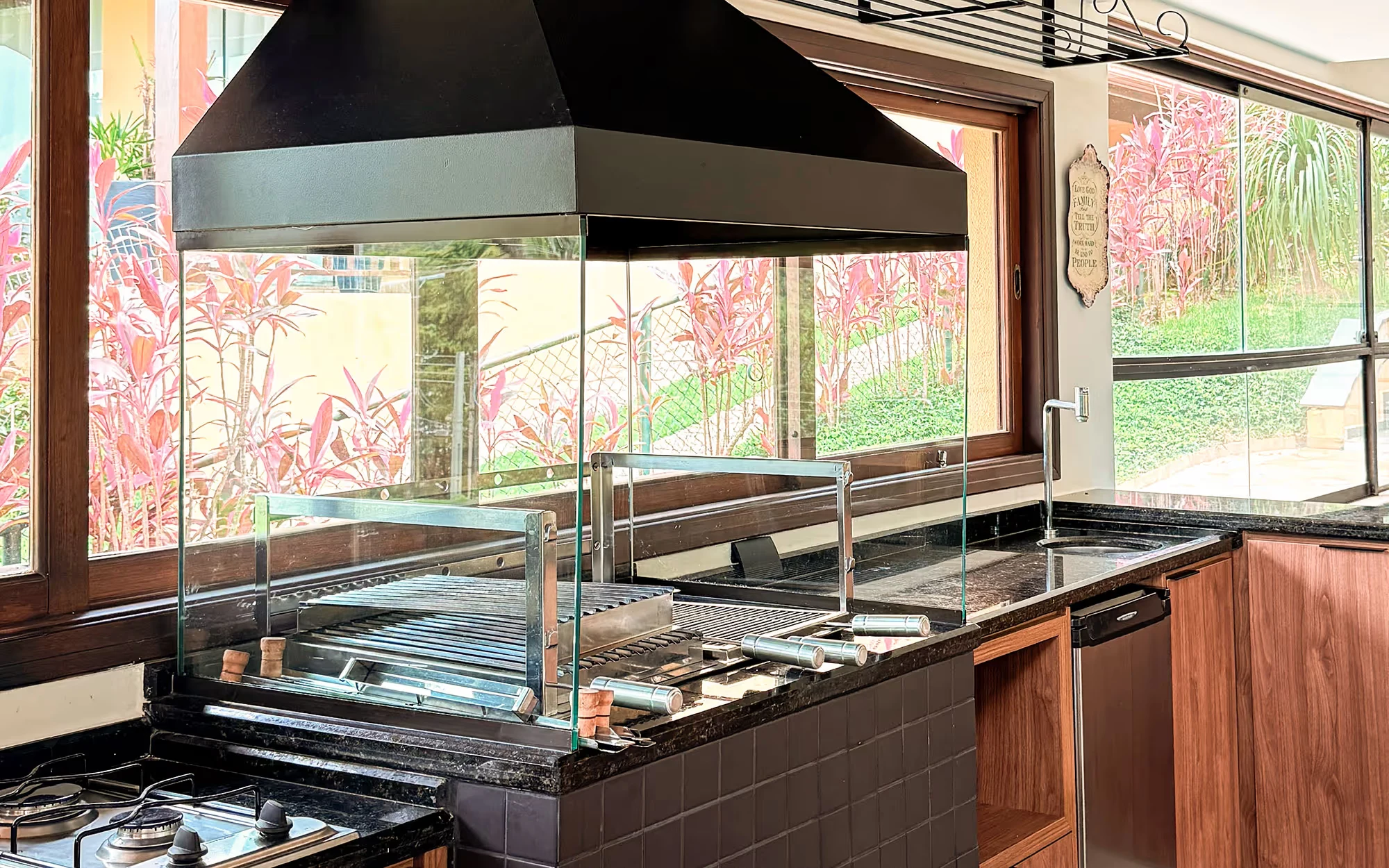 Churrasqueira com fechamento de vidro sob uma coifa preta, com bancada de tampo escuro, cooktop a gás e armários de madeira, e plantas de folha rosa lá fora., Casa Flamboyant Gourmet, Valinhos, São Paulo