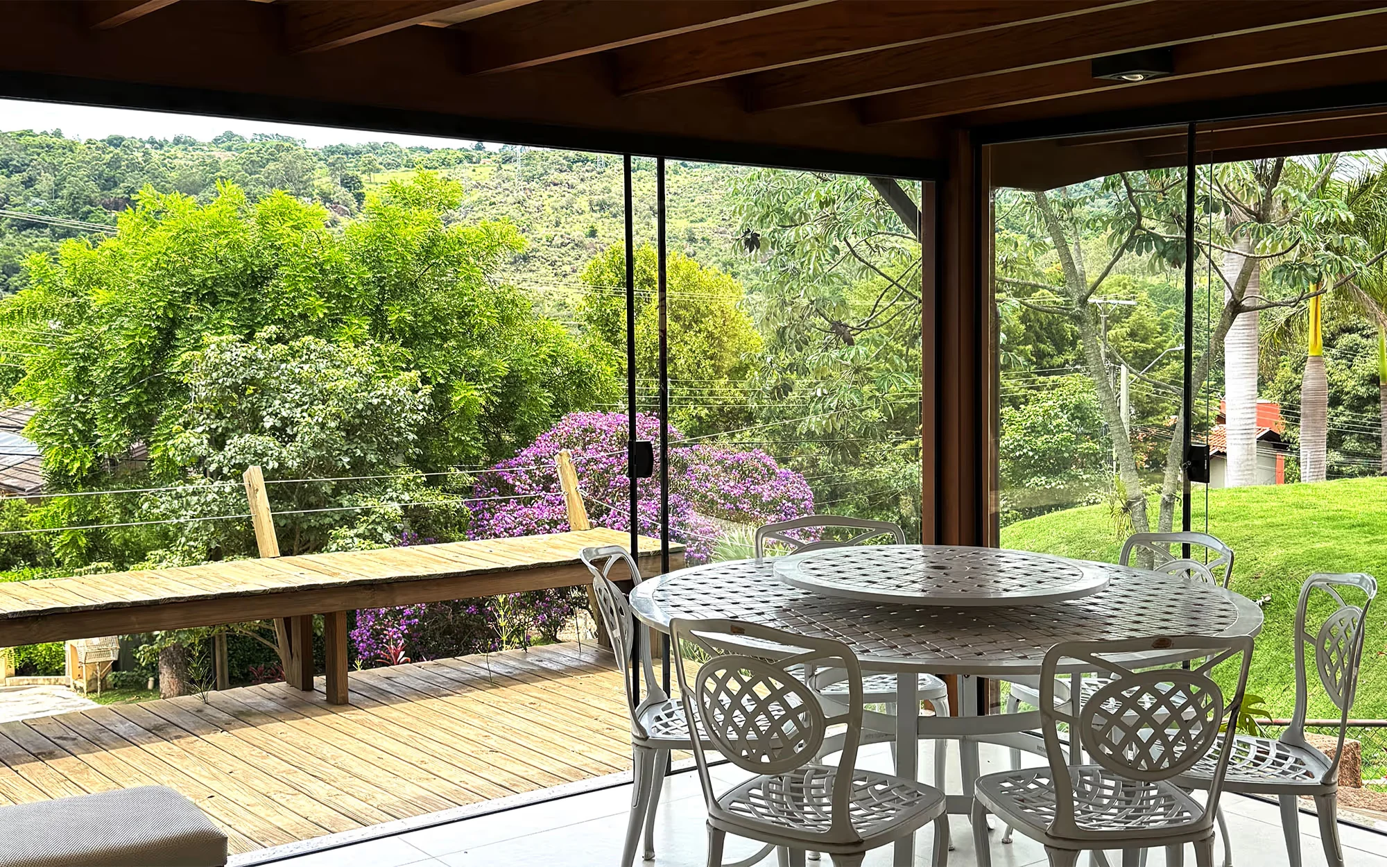 Varanda envidraçada com mesa redonda e cadeiras de metal branco, sobre um deck de madeira, vegetação com flores roxas e uma encosta verde., Casa Flamboyant Gourmet, Valinhos, São Paulo