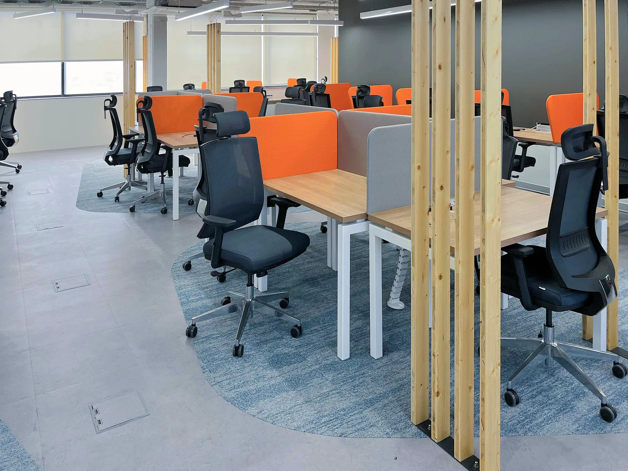 Plateau aberto com secretárias bench de tampo carvalho e estrutura branca, divisórias acústicas laranja e cinzentas, e tecto técnico à vista com iluminação linear LED suspensa.