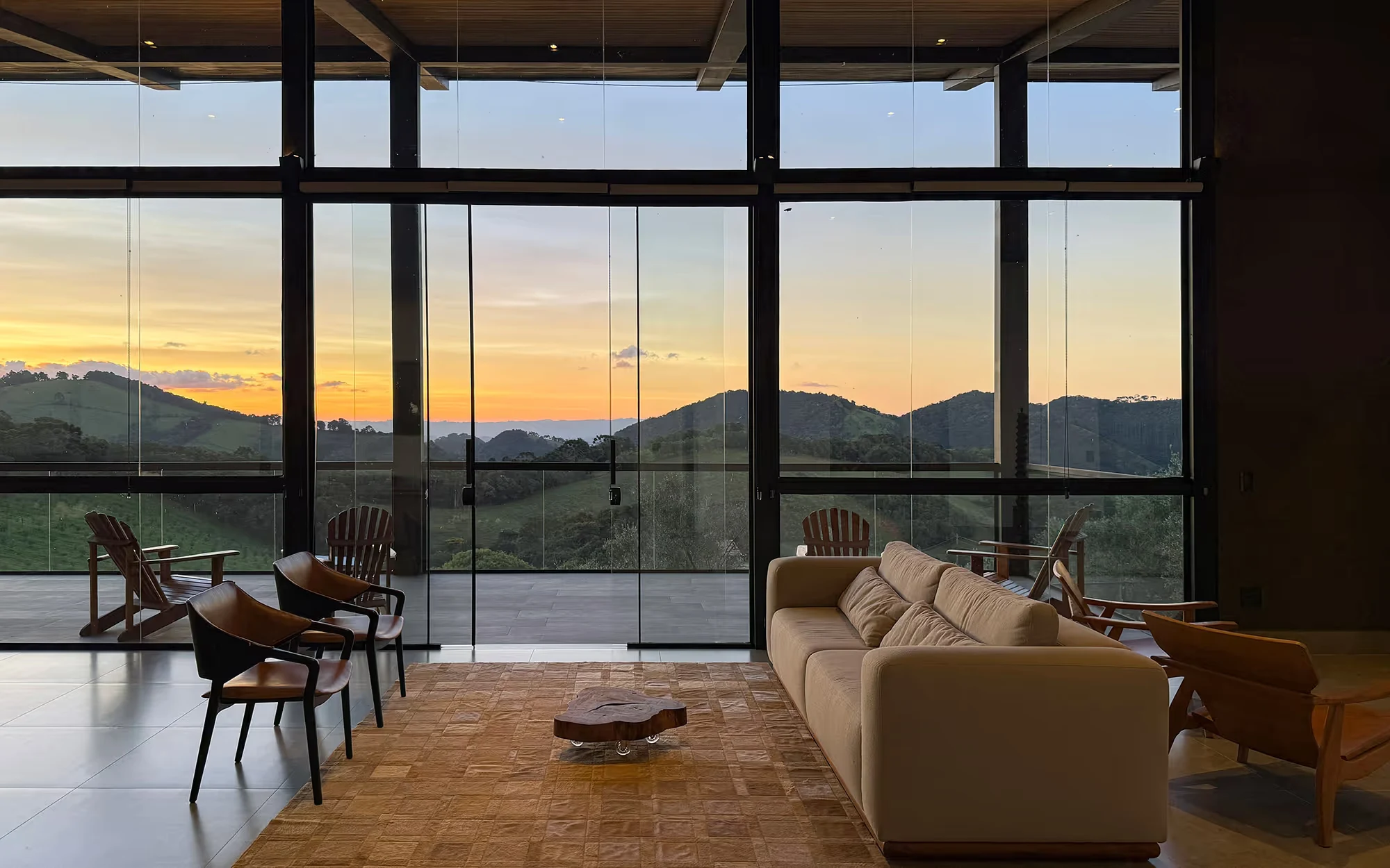 Com o céu alaranjado, sala com vãos envidraçados do piso ao teto voltados para uma serra, com sofá claro, cadeiras e um tapete, e cadeiras de madeira no terraço., Lagar Pura Oliva Brasil, São Mateus de Minas, Minas Gerais