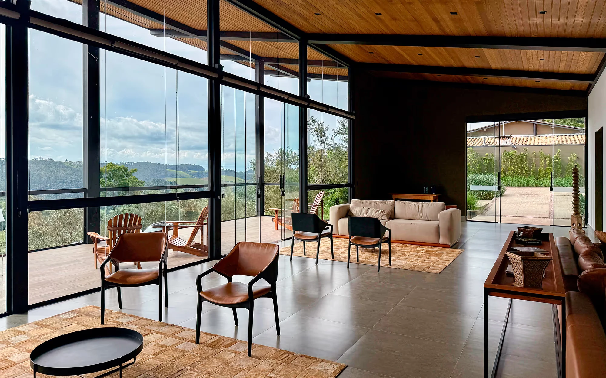 Sala ampla com parede envidraçada de esquadrias escuras e forro de ripas, cadeiras, sofás e tapetes, e uma porta envidraçada aberta para um pátio ao fundo., Lagar Pura Oliva Brasil, São Mateus de Minas, Minas Gerais