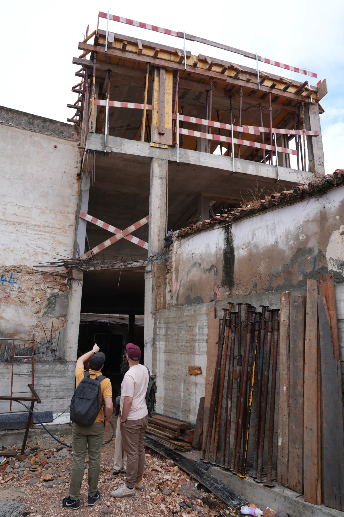 Um muro de rua de pé sozinho, com uma estrutura de betão a subir por trás e por cima dele; duas pessoas ao pé a olhar para cima, e um conjunto de prumos encostado à parede.