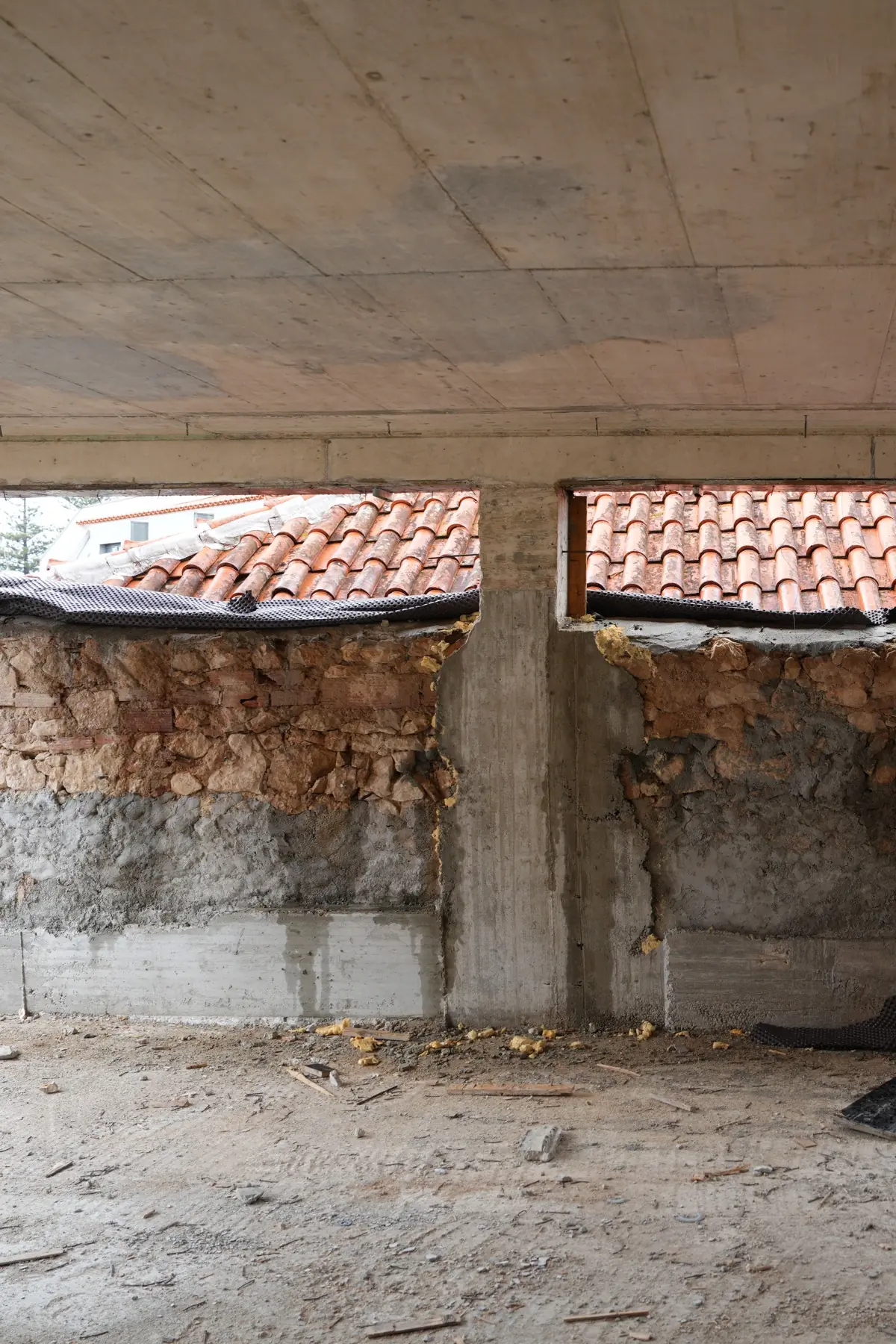 Dentro da estrutura: laje e elementos verticais de betão em cima, um muro de alvenaria de pedra em baixo, e duas aberturas longas por onde se vêem telhados de telha vizinhos.