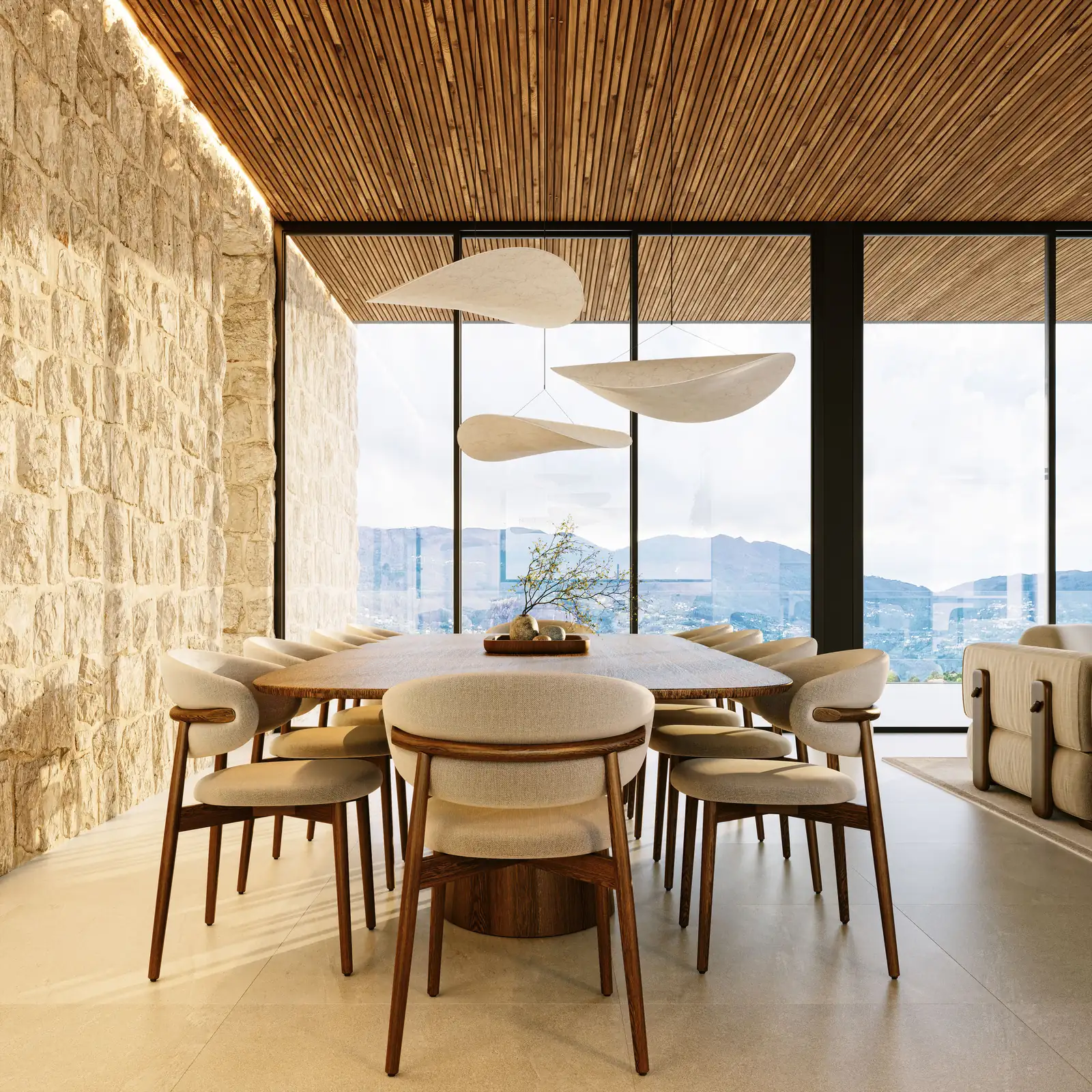 A sala de jantar contra uma parede de pedra aparelhada, mesa redonda com seis cadeiras, três candeeiros claros em forma de folha por cima e o vale pelo envidraçado à esquerda.