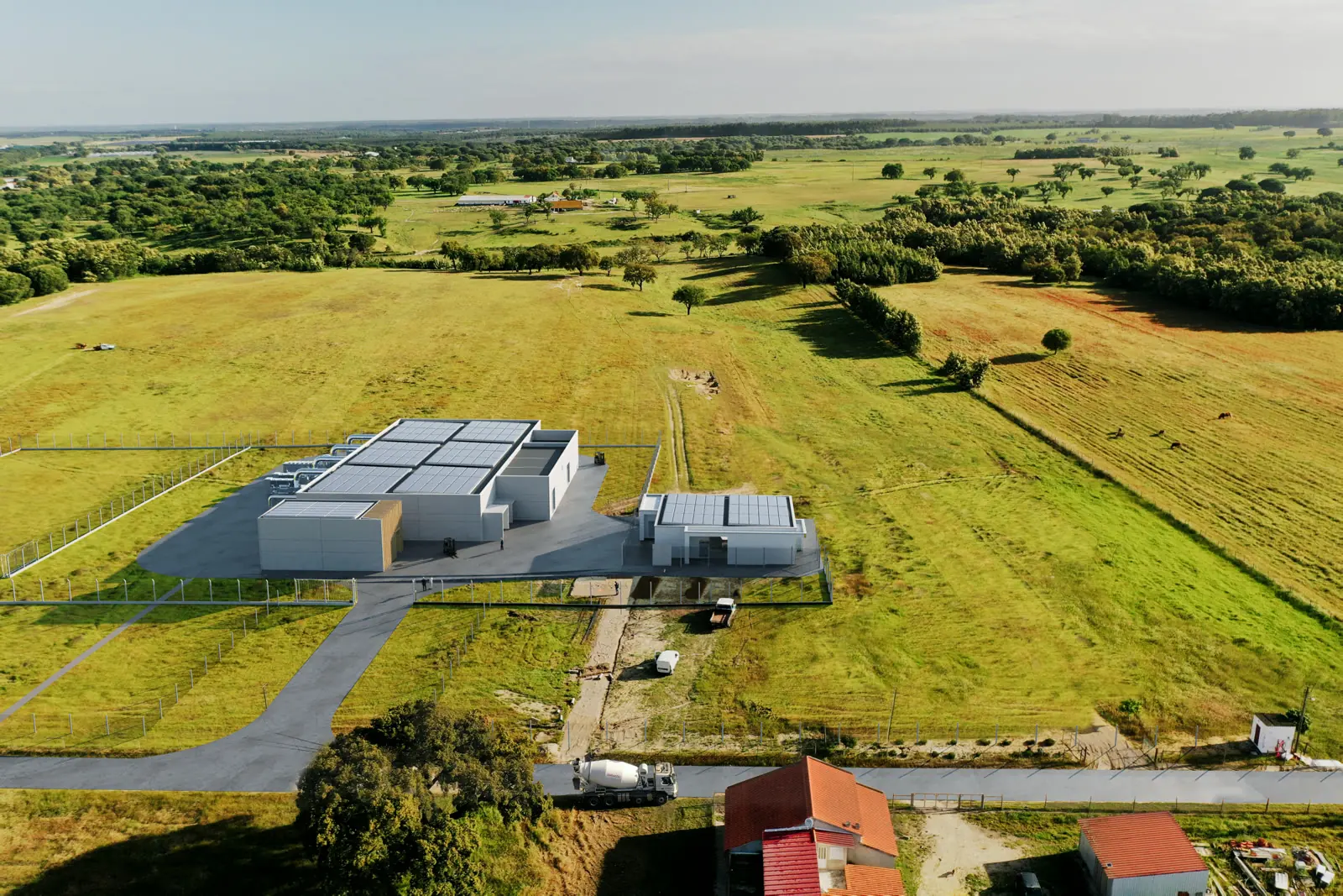 Um render aéreo do conjunto na paisagem: volumes brancos e um estaleiro pavimentado no meio de campos verdes e linhas de árvores.